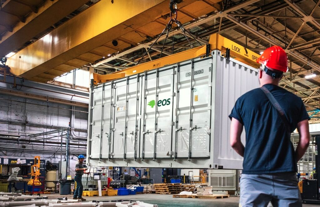 A battery storage container marked with the logo of manufacturer Eos, is lowered into place inside a factory or warehouse. 