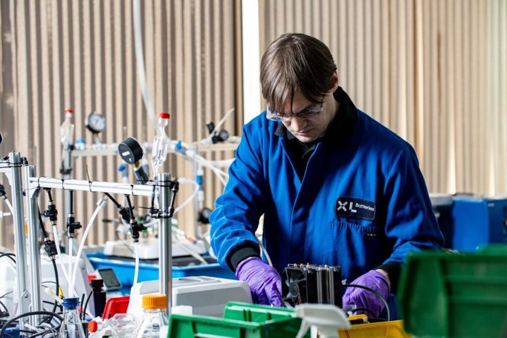 A worker in a blue branded lab coat surrounded by scientific equipment at XL Batteries' laboratory facility.