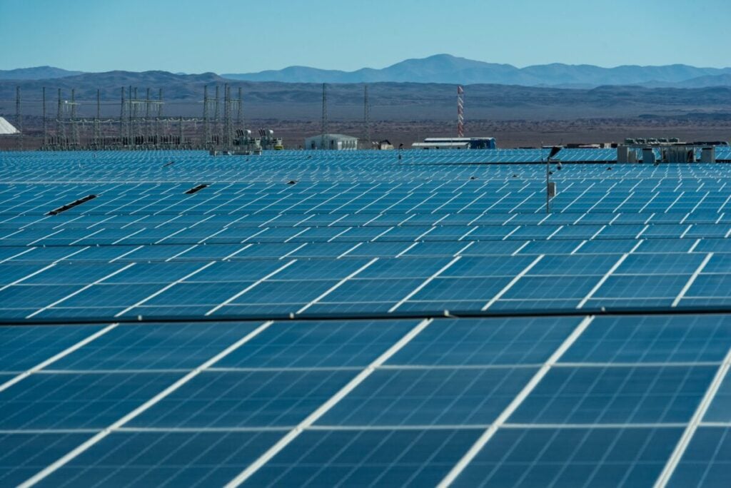 Array of dark blue photovoltaic modules in Chile's Atacama Desert, with electrical substation infrastructure in front of mountains in background.