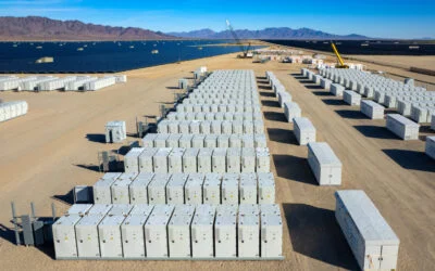 Aerial view of industrial battery units storing electricity in the desert. In the distance are solar panels and mountains.